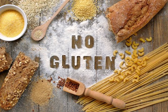 Gluten free flour and cereals millet, quinoa, corn flour polenta, brown buckwheat, basmati rice and pasta with text gluten free in English language with wooden spoon on brown wooden background