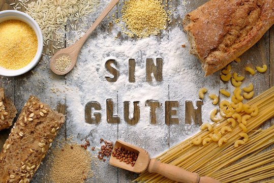 Gluten free flour and cereals millet, quinoa, corn flour polenta, brown buckwheat, basmati rice and pasta with text gluten free in Spanish language with wooden spoon on brown wooden background