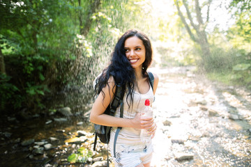 Outdoor shot of attractive young woman with backpack standing in a mountain stream. Female hiker in creek water.Portrait of young woman holding bottle of water. Happy woman enjoying tropical rain