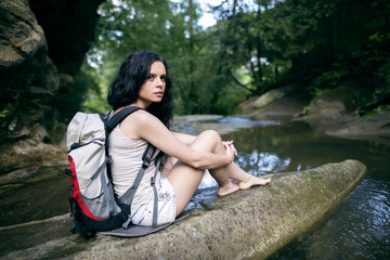 Portrait of beautiful young woman with backpack sitting on the rock. Female hiker hiking by the mountain stream.