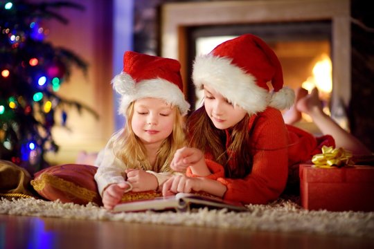 Two cute little sisters reading a story book together under a Christmas tree