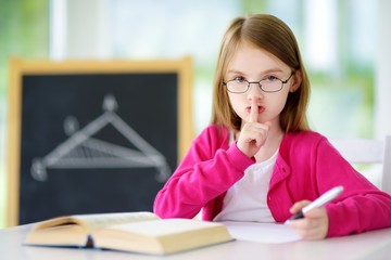 Smart little schoolgirl with pen and books writing a test in a classroom