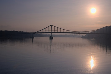 Fototapeta na wymiar Beautiful morning cityscape. The sun is reflected in water. silhouettes of houses hidden in the haze. Pedestrian bridge to the Trukhanov island