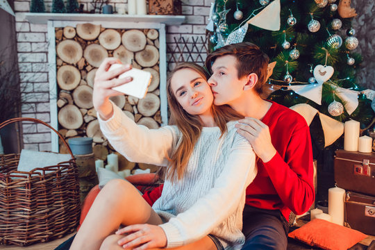 Young couple in love sitting near christmas tree having fun, kiss and make selfie. New Year