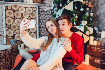 Young couple in love sitting near christmas tree having fun and make selfie. New Year