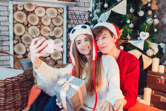 Young couple in love sitting near christmas tree in funny hats having fun and make selfie. New Year