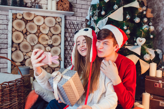 Young couple in love sitting near christmas tree in funny hats having fun and make selfie. New Year