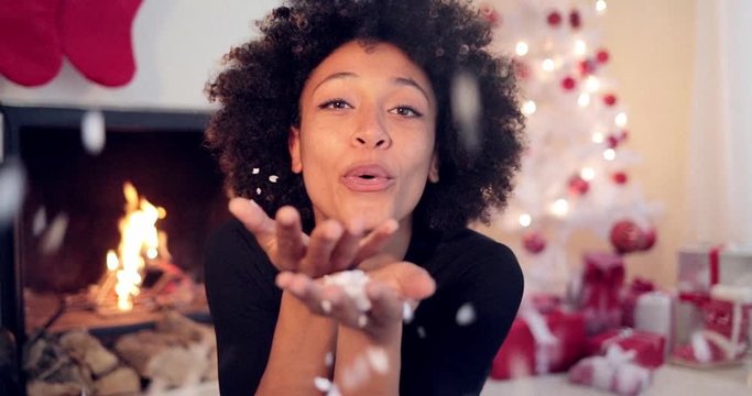Young african american woman with big afro haircut blowing white confetti off her hands as she celebrates Christmas at home in front of a burning fireplace and beautifully decorated white Xmas tree.