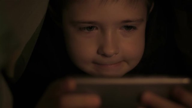 Six year old boy playing on tablet under covers. Close up. Little boy lying under covers at night. Teenager under covers playing on tablet and reading book