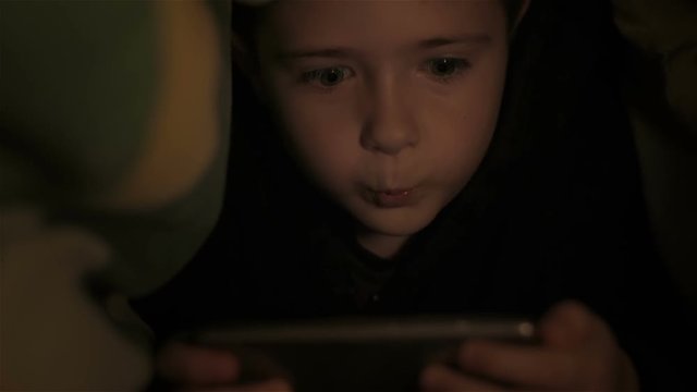 Boy playing on tablet under covers and wonders. Close up. Little boy lying under covers at night. Teenager under covers playing on tablet and reading book