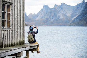 Man photographing mountains, sitting on a wooden pier. Makes selfie