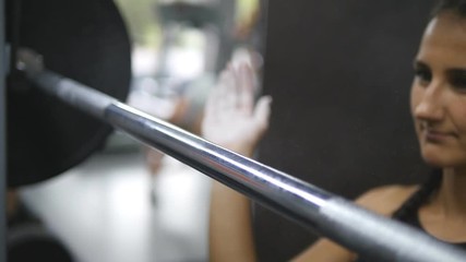Portrait of young woman prepares to lift heavy barbells at the gym. Taped hands of girl clapping white chalk powder. Female athlete taking a barbell with heavy weights on her shoulders. Slow motion