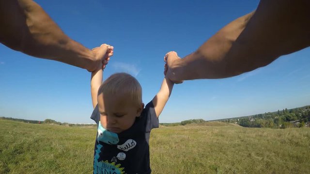 Father Spinning His Little Son In A Meadow On A Sunny Day. Dad Rotate Cute Baby Around At Nature. Boy Hold Hands Of Parent. Family Spends Time Together Outdoor. POV - Point Of View. Close Up