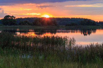The sun sets behind the forest on a dark lake, summer evening, horizontal frame
