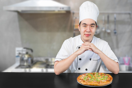 Asian young man chief in white uniform offers to you delicious pizza.