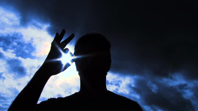 Person silhouetted raises hand into frame to give an a-okay sign with sun shining through to camera lens.