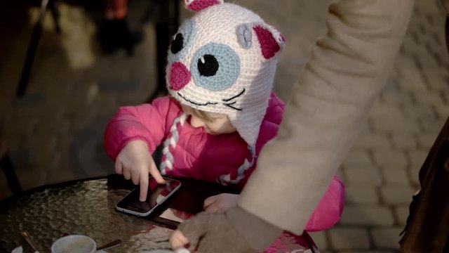 Pretty Little Girl Using App on Smartphone Sits in the Street Terrace