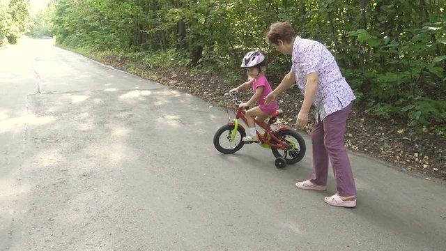 Grandmother learns to ride a bike little granddaughter. Happy child riding a bike in park outdoor.