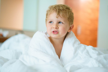 Thoughtful little blond curly haired kid looking somewhere to the side. Sitting up in bed, wrapped in a white blanket. Blurry background.