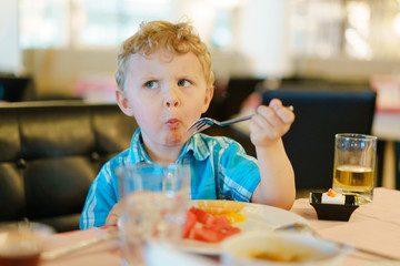 Blond kid tries breakfast fruits with suspicious wary expression on his face. He keeps fork in his...