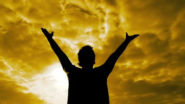 Silhouette of person waling towards camera, raises his arms and hands to the vivid yellow sunny sky.