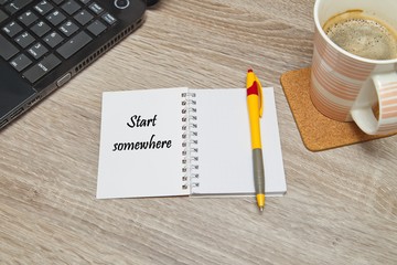 Open notebook with Text "Start Somewhere" and a cup of coffee on wooden background. Top down viewffice