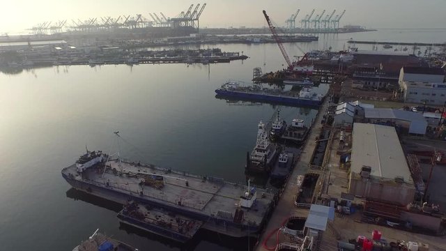 Aerial shot over the industrial port harbor in Long Beach, California.  Ships, barges, cranes, and shipyard visible early in the morning with sun on the left. Still, calm water.