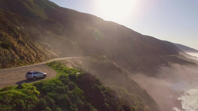 Aerial revolving shot of a Subaru car parked on the side of a cliff overlooking the ocean in Big Sur, California. Dazzling sunlight with fog and mist down the coast. 