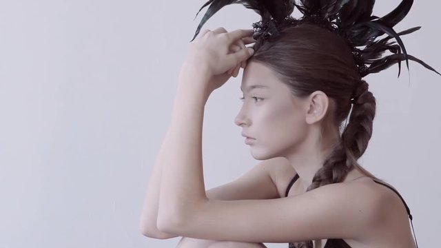 Young mixed race caucasian woman vogue portrait with feather mohawk accessory wearing black bodysuit sitting on wooden floor on white wall background. Fashion concept.