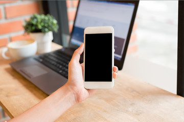 Hand holding blank screen smartphone mobile in coffee shop with laptop in background