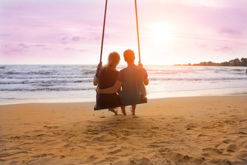 Romantic couple is sitting on sea beach on rope swing and looking at sunset horizon. Family...