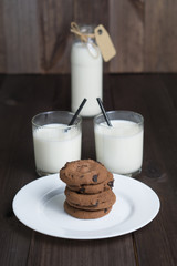 two transparent glasses full with milk with carton label tag, black straw and pile brown cookies on white plate with blurred bottle of milk on dark wooden background