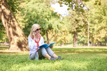 Fototapeta premium Woman using her mobile outdoor. Full length shot of a middle aged businesswoman working in the park and making call while holding in her hand some document.