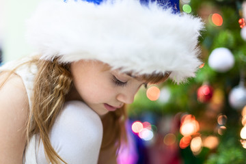 Calm little girl in fur Santa hut sitting near th Christmas tree with light, close-ip view