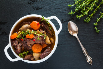 Beef Bourguignon in a white soup bowl on black stone background, top view. Stew with carrots, onions, mushrooms, bacon, garlic and bouquet garni. The dish is served with fresh thyme. - 128393236