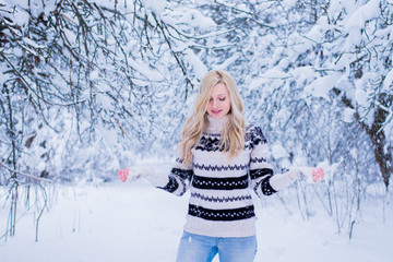 Beautiful young woman with blond hair is dressed in a warm black and white sweater on the snowy background in the winter forest. / Beautiful young woman throwing snow and smiling. Christmas theme