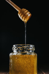 Crop hand dipping wooden honey stick in glass jar of honey isolated on black .