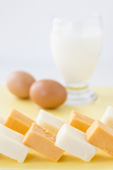 cubed blocks of white and orange cheese in forefront with blurred background of glass of milk and two brown eggs on white background