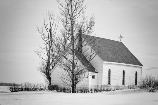 horizontal black and white image of an old rustic white wooden church sitting on a blanket of snow in the winter time.