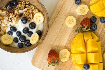 brunch : home made yogurt with granola and banana in wooden bowl. The mango cut on a dnrnvyany board and decorated with strawberry, blueberry and bananas.mango on a dark wood background. tinting.


