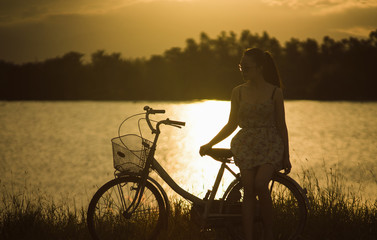 teen asian woman sit at retro vintage bicycle near the lake at sunset moment. silhouette bicycle at the sunset with grass field.big mountain and sunset background.journey concept.lonely concept