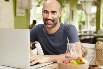 Fototapeta premium People, lifestyle and modern technology. Outdoor portrait of happy handsome man sitting at cafe table with glass of smoothie and food during breakfast, keeping hands on keyboard of his generic laptop