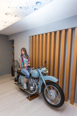 Fototapeta na wymiar Cool biker woman in grey t-shirt and blue jeans sitting on old fashioned motorcycle in garage interior on wooden wall background, vertical picture