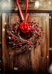 Handmade Christmas wreath hanging on aged old wooden door with red ribbon and berries with snow, ...