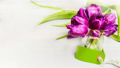 Tulips bunch in glass jar with water and empty tag on light background, banner. Spring flowers concept.