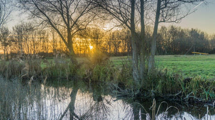 Blick auf Wiese, Baeume und  Sonnenuntergang über einem See, mit orangefarbenen Abendrot