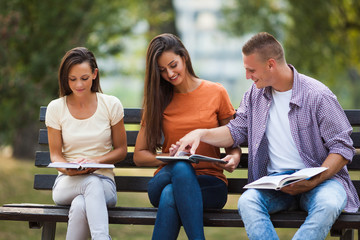 Three students are sitting on bench in park and learning. 