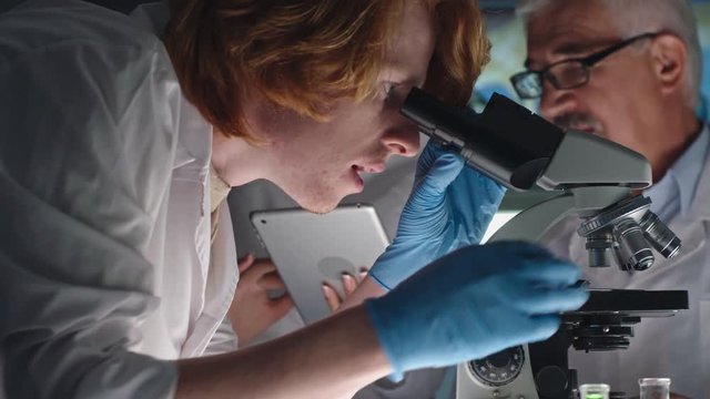 Tracking shot of young student looking through microscope while woman and senior scientist discussing something on tablet screen