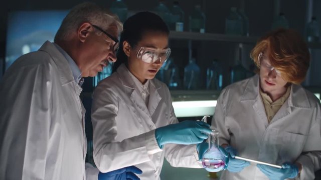 Female chemist mixing substances in flask and looking at reaction while young student using tablet and mature professor talking to her