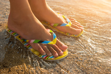 Female feet wearing flip flops. Water and sand under sunlight. One step away from sea. Visit a...